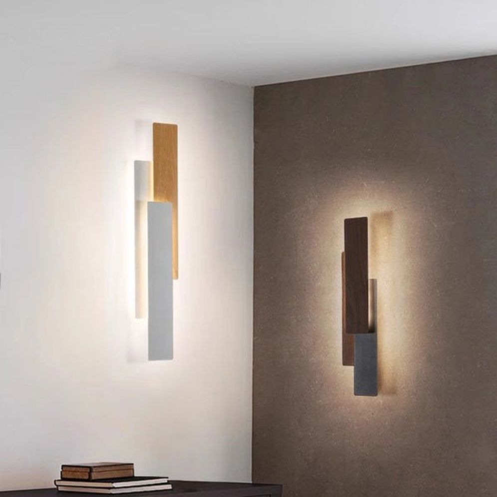 Two Applique murale bois LED rectangulaire design moderne wall sconces, with wood and metallic finishes, are mounted on adjacent walls to provide a warm, diffuse ambient light; a desk with several books sits in the foreground.