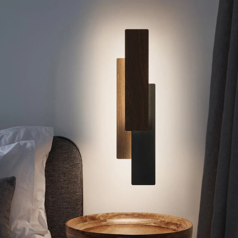 The Applique murale bois LED rectangulaire design moderne creates a warm, diffuse glow with overlapping wood and black panels, beautifully illuminating a round wooden bedside table beside a gray upholstered bed with white pillows.