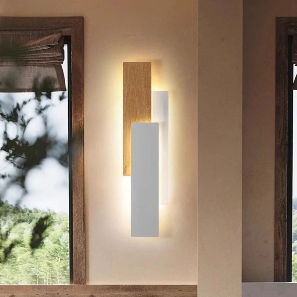 The Applique murale bois LED rectangulaire design moderne features overlapping wooden and white panels, casting a warm diffuse light on a beige wall near a window with a roman shade and green tree views.