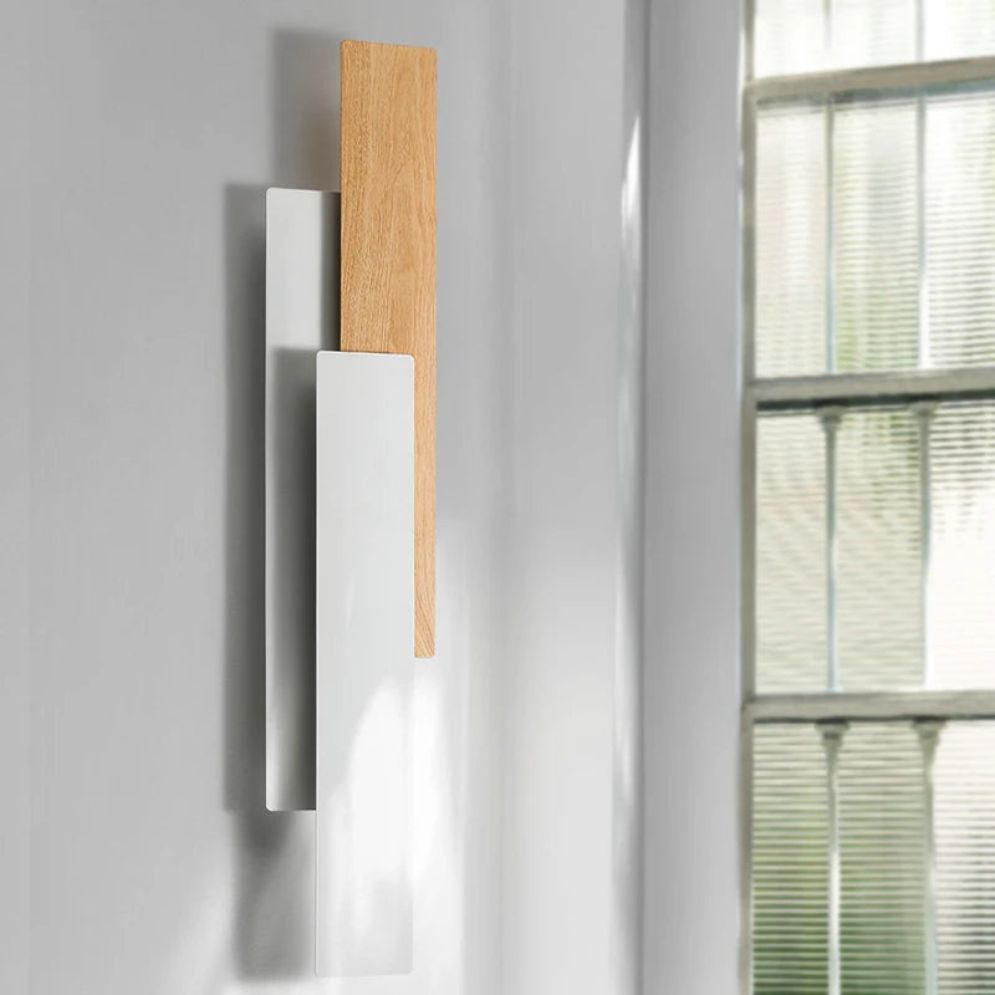 A modern Applique murale bois LED rectangulaire design features overlapping light wood and white panels, mounted on a white wall near frosted glass, creating warm ambient lighting.